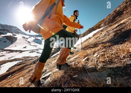 Zwei aktive Wanderer in leuchtend gelben und orangen Kleidern gehen hoch in den Bergen gegen schneebedeckte Berge Stockfoto