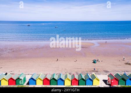 Farbenfrohe Strandhütten oberhalb von West Beach, Whitby, mit Urlaubern, die die Sommersonne am Sandstrand genießen, und Booten auf dem ruhigen blauen Meer. Stockfoto