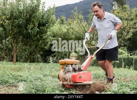 Ein reifer Mann fokussiert sich intensiv, während er mit einer Motorhacke den Boden in seinem Garten für das Pflanzen vorbereitet. Das Bild zeigt die Inanspruchnahme und die harte Arbeit Stockfoto