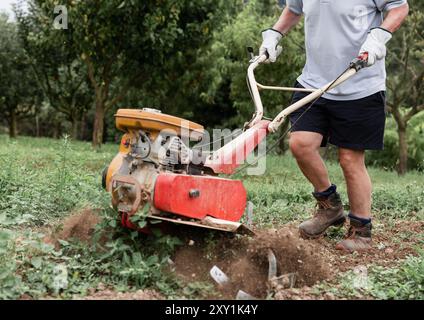 Eine Nahaufnahme eines Mannes, der mit einer Bodenfräse den Boden in seinem Garten zum Pflanzen vorbereitet. Das Bild konzentriert sich auf die harte Arbeit und Präzision Stockfoto