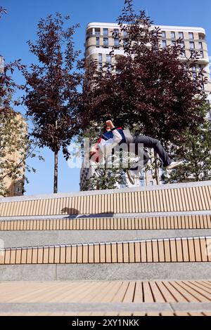 Der junge Mann im Hintergrund, dessen Silhouette sich von hohen Bäumen und urbaner Architektur abhebt, verkörpert den Nervenkitzel dynamischer Bewegungen in der Stadt. Stockfoto