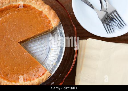 Draufsicht auf einen Feiertags-Kürbiskuchen mit einem Ausschnitt. Horizontales Format mit Servietten, Teller und Gabeln. Stockfoto