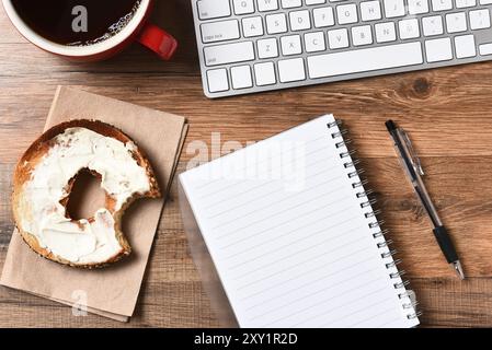 Ein Bagel mit Frischkäse und einem Biss, der neben einer Computertastatur und einem Pad und Stift herausgenommen wurde. Eine Tasse Kaffee in der oberen Ecke, Horizontal für einen Hi Stockfoto