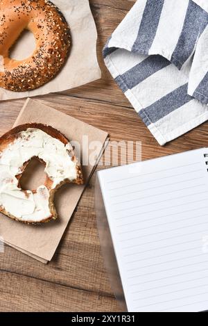 Ein Bagel mit Frischkäse und einem Biss. Mit Handtuch und Notizblock auf einem rustikalen Holzschreibtisch. Hochwinkelansicht im vertikalen Format, Stockfoto