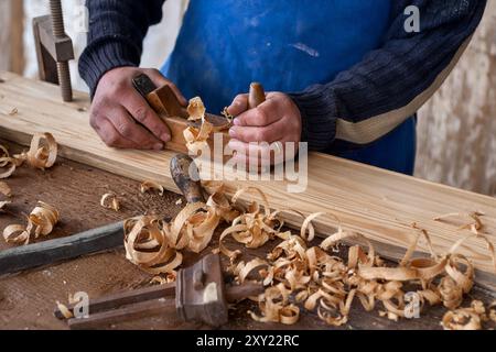 Nahaufnahme eines Handwerkers, der in einer traditionellen Werkstatt ein Holzbrett mit gelockten Holzspänen von Hand hobelt Stockfoto