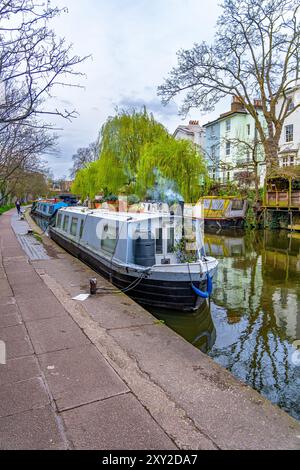 Frau, die entlang der Regent's Canal Promenade in Londons Little Venice mit farbenfrohen schmalen Hausbooten läuft, die am Fluss in der Residenz angedockt und vertäut sind Stockfoto