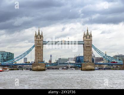 Vorderansicht der Londoner Tower Bridge von der Themse mit Straßenverkehr auf der Straße und Fähren, die unter bewölktem Himmel entlang des Flusses segeln. Vereint Stockfoto