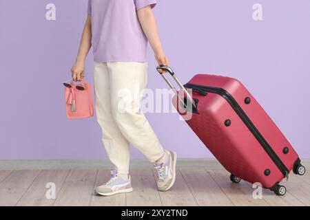 Junge Frau mit Koffer, Handtasche und Sonnenbrille auf Fliederhintergrund Stockfoto