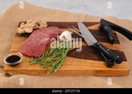 Rohes Rindersteak mit Ingwerwurzel, Knoblauch, Sojasauce und Rosmarin auf einem Holzbrett auf dem Küchentisch Stockfoto