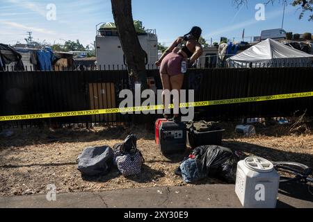 Sacramento, CA, USA. August 2024. Marie Mowder, eine zweijährige Bewohnerin von Camp Resolution, steht auf ihrem tragbaren Generator außerhalb des Zauns, als Dutzende von Polizeibeamten von Sacramento, der Abteilung für Gemeinschaftsreaktion, öffentliche Arbeiten, die Durchsetzung von Kodizes, die Tierkontrolle und die Feuerwehr am Montagmorgen die Lagerauflösung ausräumen, nachdem sich die Bewohner des selbstverwalteten Obdachlosen-Lagers weigerten, zu verlassen und sich am Montag, 26. August 2024 in Sacramento verbarrikadiert haben. Die Frist für eine Gruppe Obdachloser, ein städtisches Grundstück namens Camp Resolution zu räumen, ist abgelaufen. Stockfoto