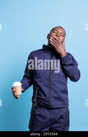 Der afroamerikanische Sicherheitsbeamte fühlt sich müde und gähnt nach der Nachtschicht und trinkt eine Tasse Kaffee, um mit Müdigkeit umzugehen. Junger Mann, der als Leibwächter arbeitet, Streifendienst und Verantwortung. Stockfoto
