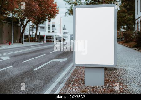 Ein leeres vertikales Plakat mit einem leeren weißen Bildschirm steht auf einem Kopfsteinpflaster neben einer ruhigen, verkehrsfreien Stadtstraße, umgeben von Bäumen Stockfoto