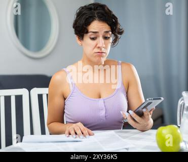 Schockierte junge Frau, die Dokumente ansieht und Smartphone in der Hand hält Stockfoto