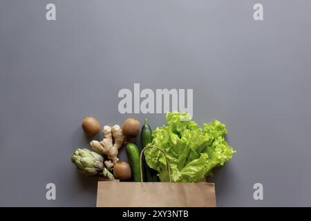 Gesundes und biologisches Lebensmittel-Flay-Lay-Konzept auf grauem Hintergrund. Wiederverwendbarer Öko-Beutel mit Salatblättern, Kiwi, Gurke, Artischocke und Ingwerwurzel Stockfoto