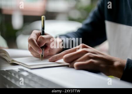Ein zugeschnittenes Bild eines männlichen College-Studenten, der in sein Notizbuch schreibt oder Notizen macht, während er online auf seinem Laptop studiert und an einem Tisch in einem Café sitzt. Stockfoto