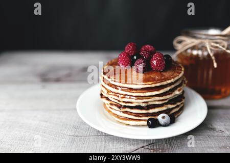 Saftige Pfannkuchen mit Beeren und Honig auf einem weißen Teller, Löffel, Glas, Holztisch. Stockfoto