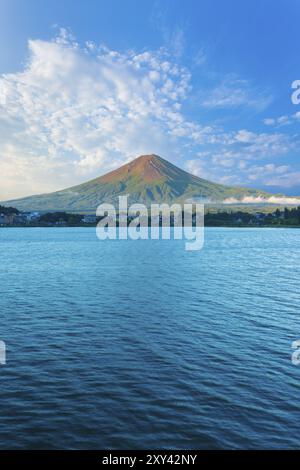 Der mittige Fuji taucht in das Morgenlicht, während der Kawaguchiko-See an einem Sommertag in der fünf-Seen-Region von J im Schatten unter dem bewölkten blauen Himmel liegt Stockfoto