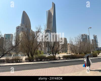 Kuwait City, Kuwait. August 2024. Eine Frau spaziert an verwelkten Bäumen vor den Wolkenkratzern von Kuwait City. Kuwait gilt als einer der heißesten Orte der Erde mit Temperaturen von über 50 °C. Quelle: Johannes Sadek/dpa/Alamy Live News Stockfoto