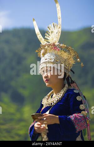 Xijiang, China, 15. September 2007: Ethnische Miao-Frau in traditionellem Festivalkostüm und silbernem Hornkopf, der eine Schüssel gegen Berg B hält Stockfoto