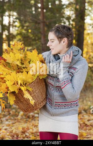 Eine junge Frau, die einen Korb voller Herbstblätter in herbstlicher, farbenfroher Umgebung trägt. Attraktive Frau sammelt Herbstblätter in einem Korb Stockfoto