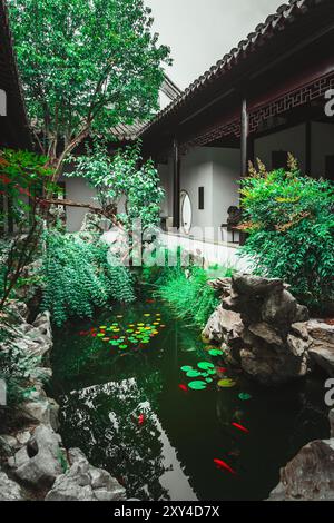 Ein kleiner Teich mit Lilienpads und rotem Fisch, eingebettet in einen traditionellen chinesischen Garten, umgeben von Stein und üppigem Grün. Stockfoto