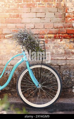 Helles, blaues Vintage-Fahrrad mit Lavendelbouquet im Korb, das gegen die schäbige Ziegelwand auf der Straße platziert ist Stockfoto