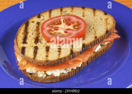 Sandwich mit Räucherlachs und russischem Salat, dekoriert mit einer Tomatenscheibe, serviert auf einem blauen Teller auf orangefarbenem Hintergrund Stockfoto