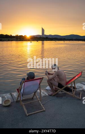 Zwei junge Männer sitzen in Liegestühlen am Ufer der Neuen Donau am Copa Beach bei Sonnenuntergang, Wien, Österreich Stockfoto