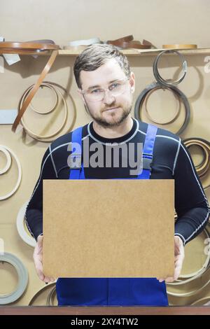 Der Hersteller von Möbeln in Spezialkleidung und Schutzbrille hält am Arbeitsplatz eine leere Platte für den Text. Handwerkliche Herstellung von Möbeln Stockfoto