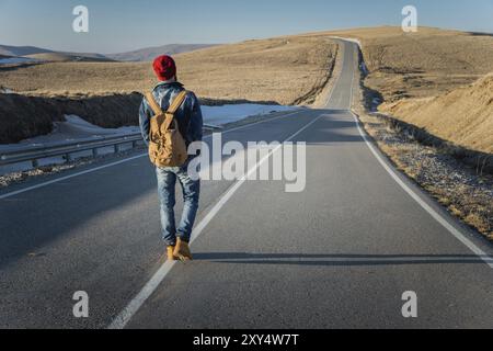 Ein bärtiger Hipster mit einem altmodischen Vintage-Rucksack, der Sonnenbrille mit rotem Hut und Jeansjacke trägt, und Jeans geht mit dem Rücken Stockfoto