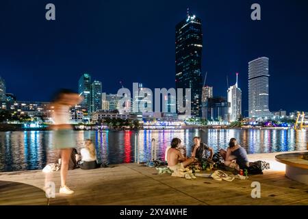 Junge Menschen, die auf einer Holzterrasse am Ufer der Neuen Donau vor der Skyline von Copa Beach und Hochhäusern der Donau City sitzen. Stockfoto