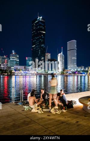 Junge Menschen, die auf einer Holzterrasse am Ufer der Neuen Donau vor der Skyline von Copa Beach und Hochhäusern der Donau City sitzen. Stockfoto