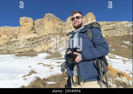 Portrait Ein bärtiger Hipster-Fotograf mit Rucksack und Sonnenbrille mit großem Rucksack auf den Schultern steht mit einer DSLR-Kamera in seinem Stockfoto