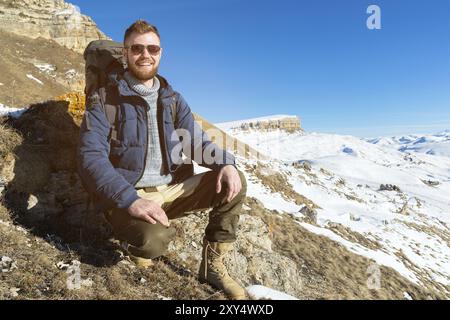 Porträt des glücklichen lachenden Hipster-Reisenden mit Bart in der Sonnenbrille sitzt auf der Natur. Ein Mann wandert in den Bergen mit Rucksack und Skandinav Stockfoto