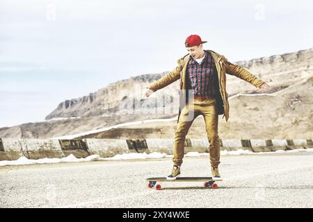 Stylischer Happy Young man in einer Mütze und Hose Jogginghose, die auf einem Longboard eine Bergstraße hinunterrollt und das Leben genießt Stockfoto