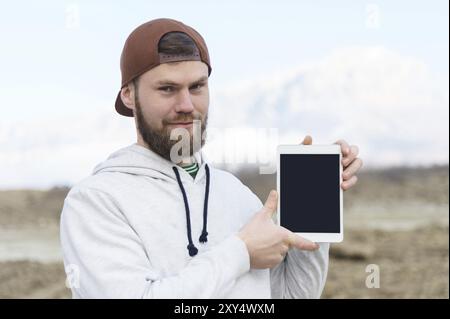 Nahaufnahme eines lächelnden Hipsters in einer braunen Kappe im Freien, der einen weißen Tablet-Computer mit einem ausgeschnittenen Rohling für den Designer hält. Eine bärtige Männerklaue Stockfoto