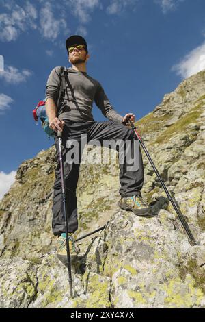 Portier eines stilvollen Hipster-Reisenden mit Bart und Rucksack in Sonnenbrille und einer Mütze mit Trekkingstöcken, die auf einem Felsen vor dem Hintergrund stehen Stockfoto