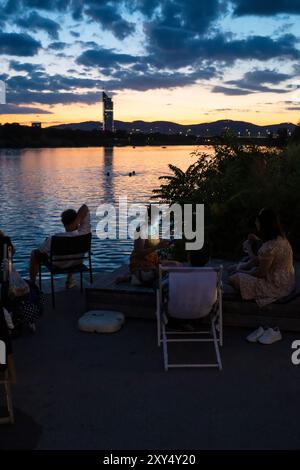 Das Gesicht einer jungen Frau am Ufer der Neuen Donau am Copa Beach während des Sonnenuntergangs wird von einem Mobiltelefon in Wien, Österreich, beleuchtet Stockfoto