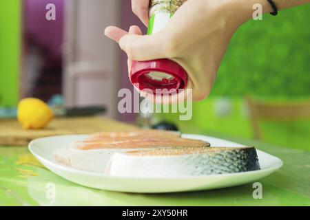 Frauenhände besprühen aus der Handmühle mit Gewürzen zwei rohe Steak vom Lachs, die auf einem weißen Teller in einer Küche in Grüntönen liegen. Das Konzept o Stockfoto