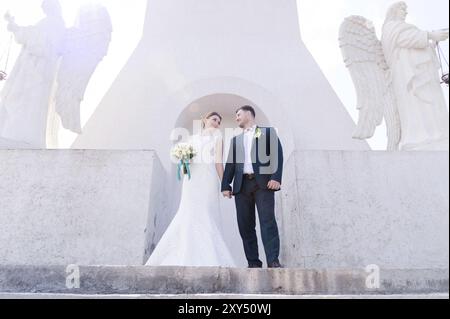 Porträt eines wunderbaren Paares, das an einem Hochzeitstag mit einem Blumenstrauß in der Hand vor dem Hintergrund eines orthodoxen christlichen Denkmals mit Engeln verbrachte. Stockfoto