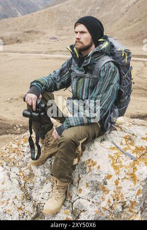 Müder bärtiger Hipster mit Fernglas in den Händen sitzt auf einem Stein zwischen den kaukasischen Bergen und blickt in die Ferne Stockfoto