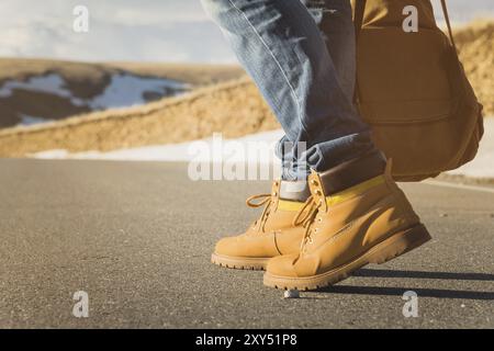 Wandern Sie auf dem Asphalt bei Sonnenuntergang im Sommer, Herrenbeine in gelben Schuhen und blaue Jeans entlang des Asphalts. Neben dem Vintage-Rucksack. Per Anhalter Stockfoto