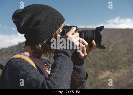 Nahaufnahme eines blonden Mädchens mit einer digitalen Spiegelreflexkamera, die in den Bergen fotografiert. Stylischer freiberuflicher Fotograf mit Rucksack in den Bergen Stockfoto