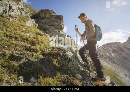 Ein bärtiger Mann mit Sonnenbrille und einer Mütze mit Rucksack steht auf einem Felsen und blickt in ein felsiges Tal hoch in den Bergen. Das Konzept der Tour Stockfoto