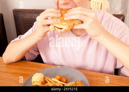 Ein Kaukasier isst halbgegessen Hamburger, Kartoffelchips und Hähnchenstücke von Hand von einem grauen Teller auf einem Holztisch, amerikanische Fast-Food-Kultur Stockfoto