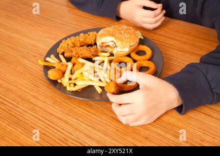 Ein Kaukasier isst halbgefressenen Hamburger, Kartoffelchips, Zwiebelringe und Hühnchenstücke von Hand von einem grauen Teller auf einem Holztisch, amerikanischer schneller Foo Stockfoto