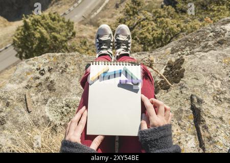 Blick von der ersten Person auf dem weiblichen Schoß ist ein Notizbuch mit Kreiden zum Zeichnen von Pastellfarben. Weibliche Hände, die ein Notizbuch in der Natur halten. Ein Mädchen sitzt Stockfoto