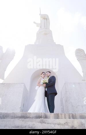 Porträt eines wunderbaren Paares, das an einem Hochzeitstag mit einem Blumenstrauß in der Hand vor dem Hintergrund eines orthodoxen christlichen Denkmals mit Engeln verbrachte. Stockfoto