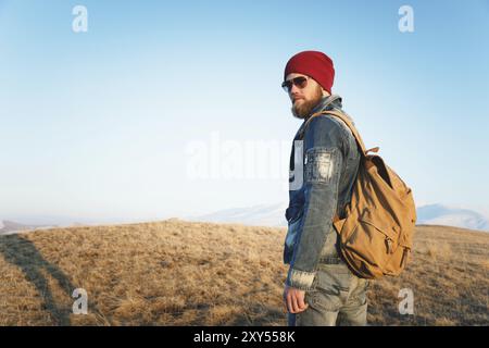 Modeporträt eines bärtigen jungen Hipster-Mannes mit Sonnenbrille, Rucksack und Hut auf einem Hintergrund mit Copyspase in den Bergen bei Sonnenuntergang, Eine Co Stockfoto