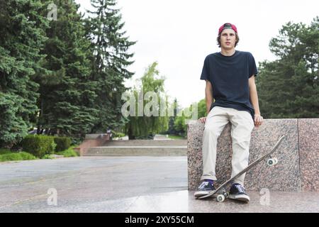 Der langhaarige Skater in Kappe und T-Shirt sitzt mit dem Rücken zur Kamera und denkt neben dem Skateboard vor dem Hintergrund der Landschaft Stockfoto